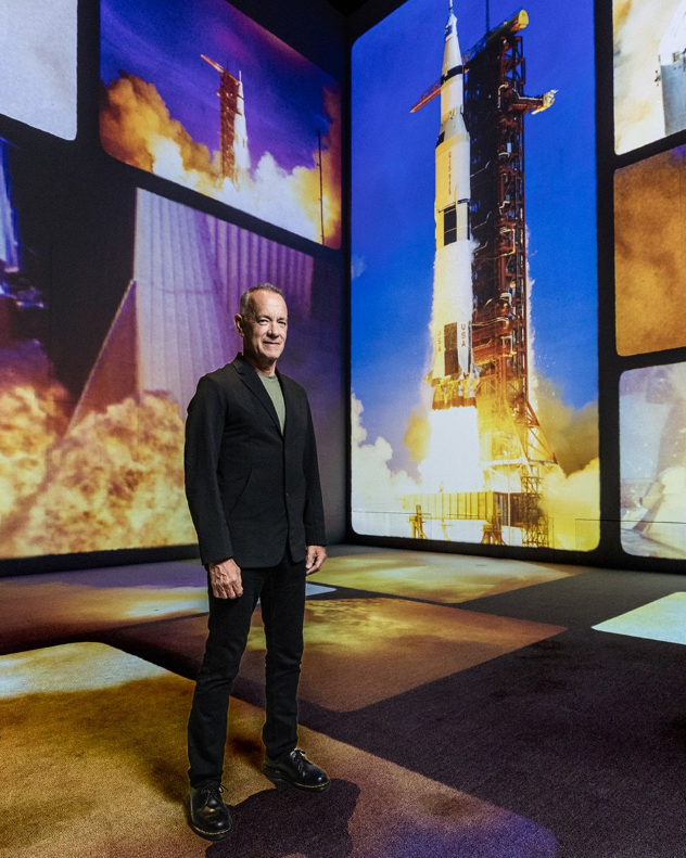 Tom Hanks stands inside The Moonwalkers immersive cinematic experience as a Saturn V rocket launches behind him